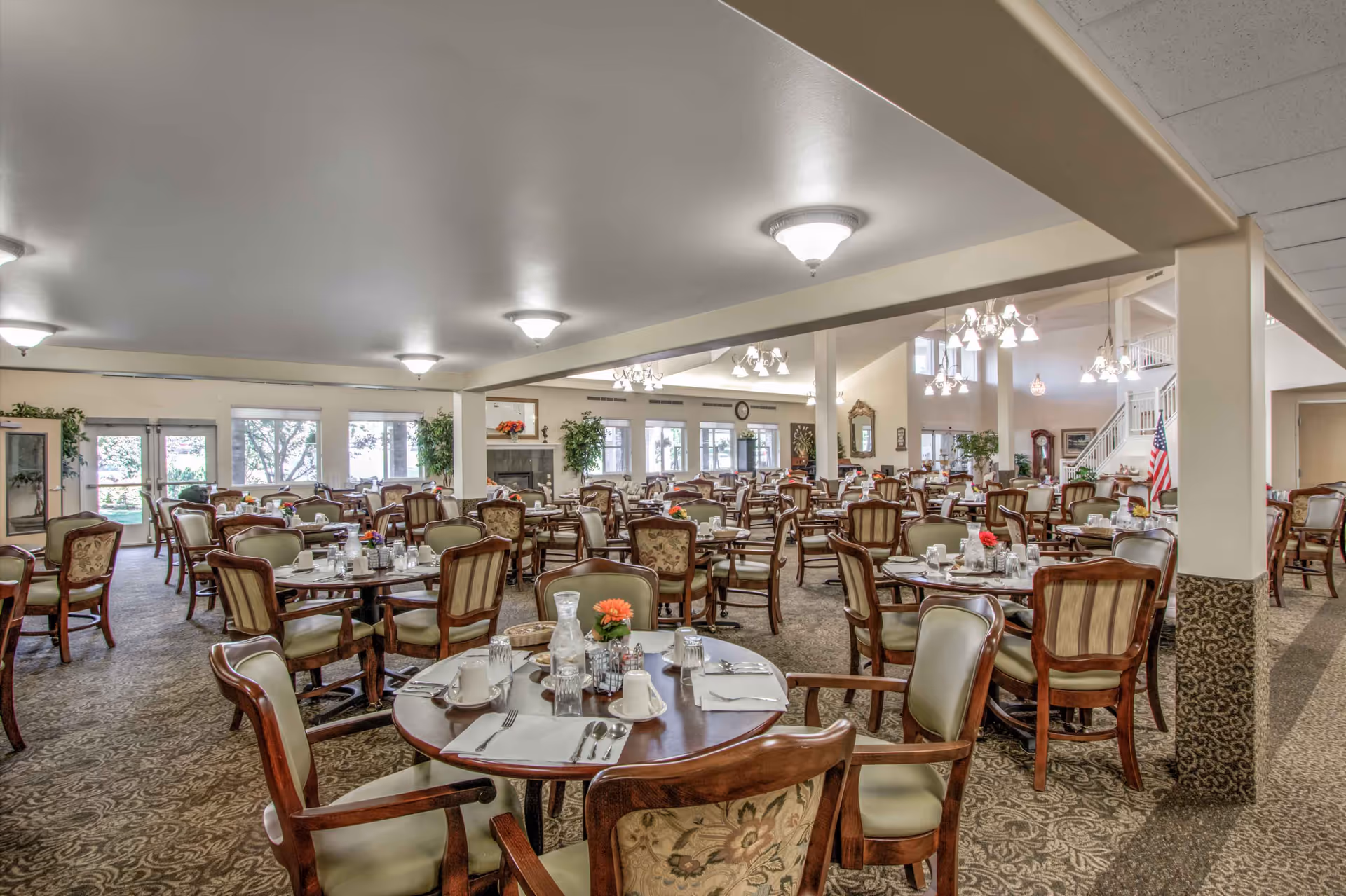 Spacious dining room with many round tables set for meals and upholstered wooden chairs.