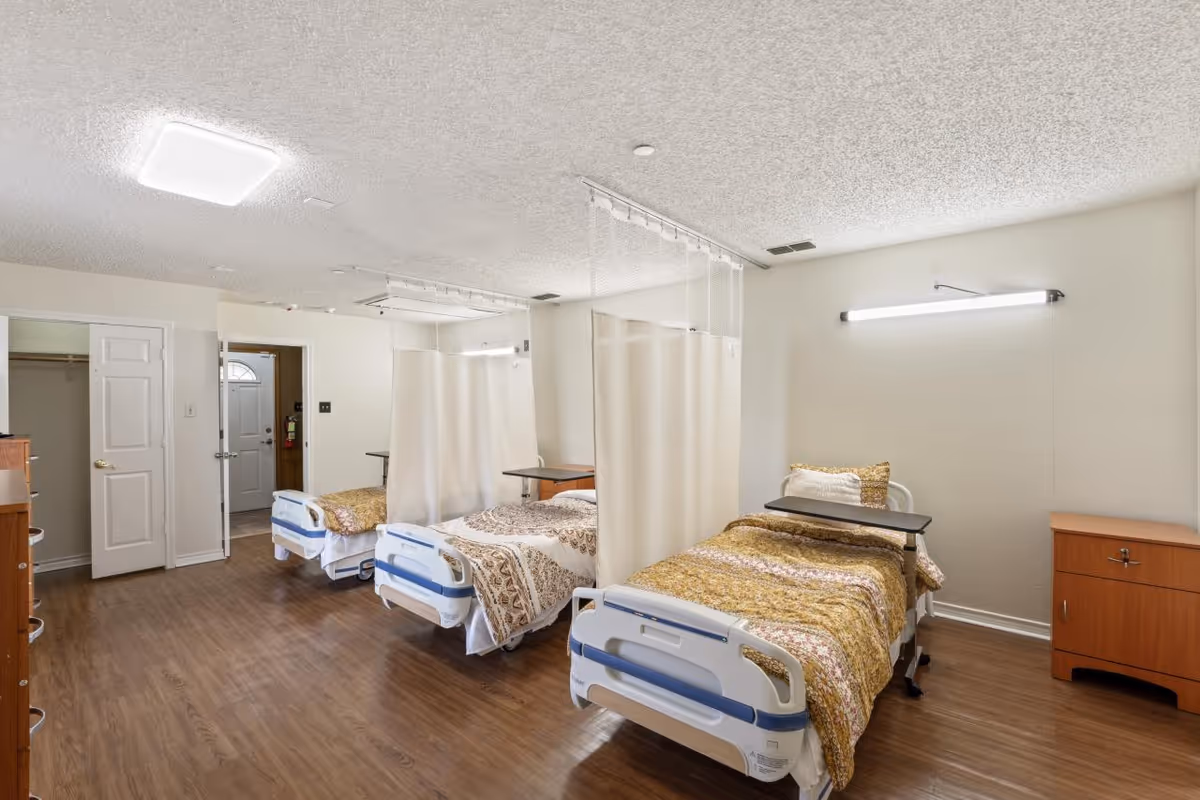 Shared assisted-living bedroom with three hospital-style beds separated by privacy curtains.
