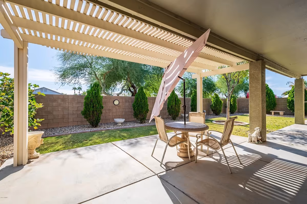 Outdoor patio area with a round table and four chairs under a pergola. A large umbrella is attached to the table. The patio overlooks a grassy yard with small trees and a brick wall fence. There are decorative stone statues near the patio columns.
