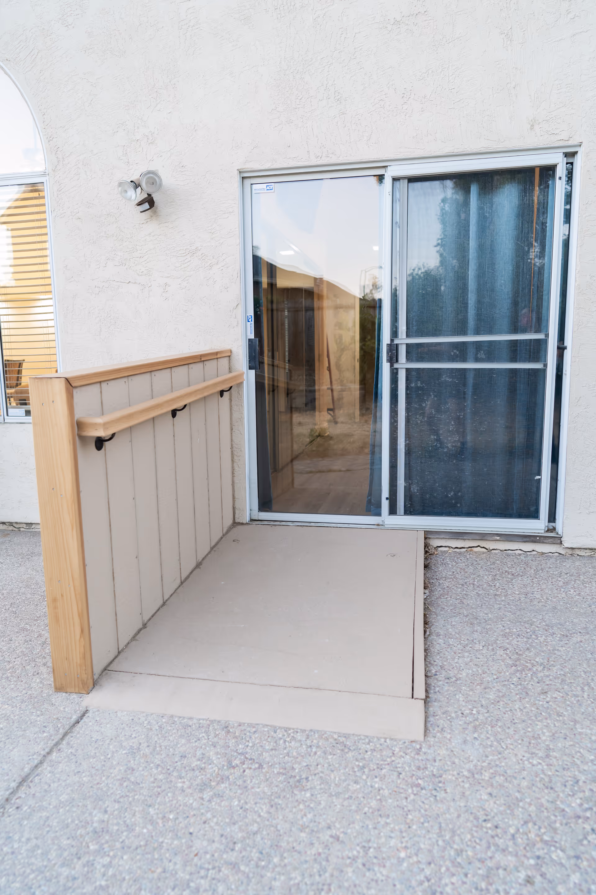 Sliding glass patio door with a small access ramp and wooden handrail against a stucco exterior wall.