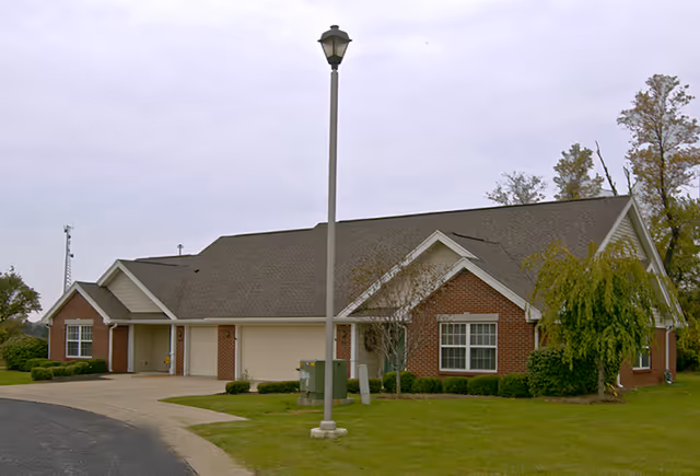 Exterior view of a single-story brick and siding building with a sloped roof, surrounded by a grassy lawn and a few trees, with a streetlamp in the foreground.
