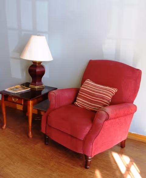 Red upholstered armchair with a striped pillow beside a wooden side table holding a lamp and a magazine against a light-colored wall.