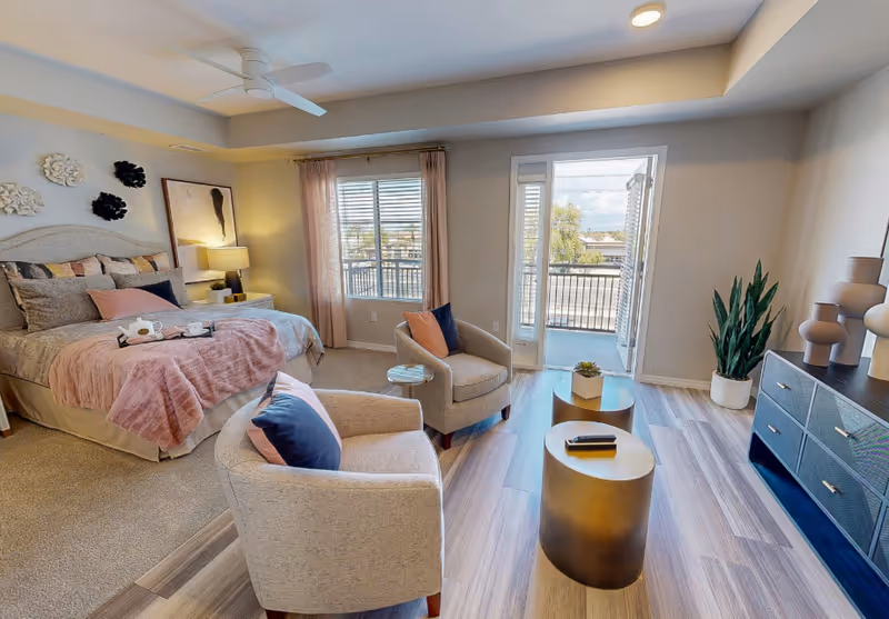 A bright and cozy bedroom with a large bed covered in pink and gray bedding, two beige armchairs with blue and pink pillows, a small round side table, and two round gold coffee tables. The room has a ceiling fan, a window with pink curtains, a glass door leading to a balcony, a potted plant, and a dark blue dresser with decorative vases on top.