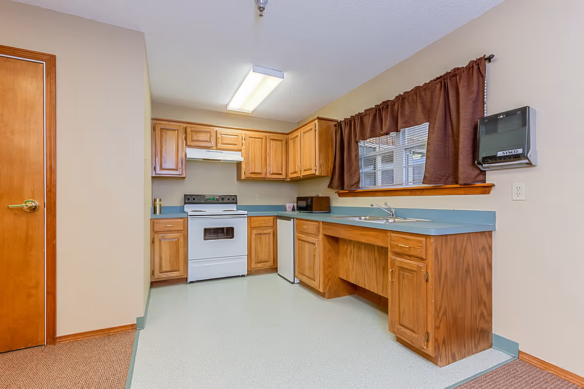 A kitchen area with wooden cabinets, a white stove, a small refrigerator, a microwave, a double sink under a window with brown curtains, and a wall-mounted Sysco device.