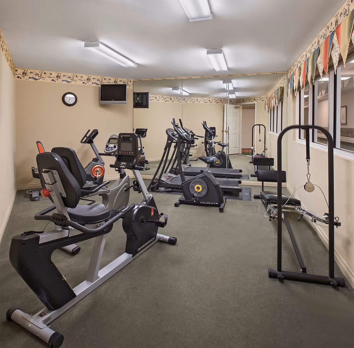 A small fitness room with exercise equipment including a recumbent bike, two upright stationary bikes, and a pulley machine. The room has beige walls, a carpeted floor, a wall clock, a mounted TV, and a large mirror covering one wall. Windows with colorful valances are on the right side.