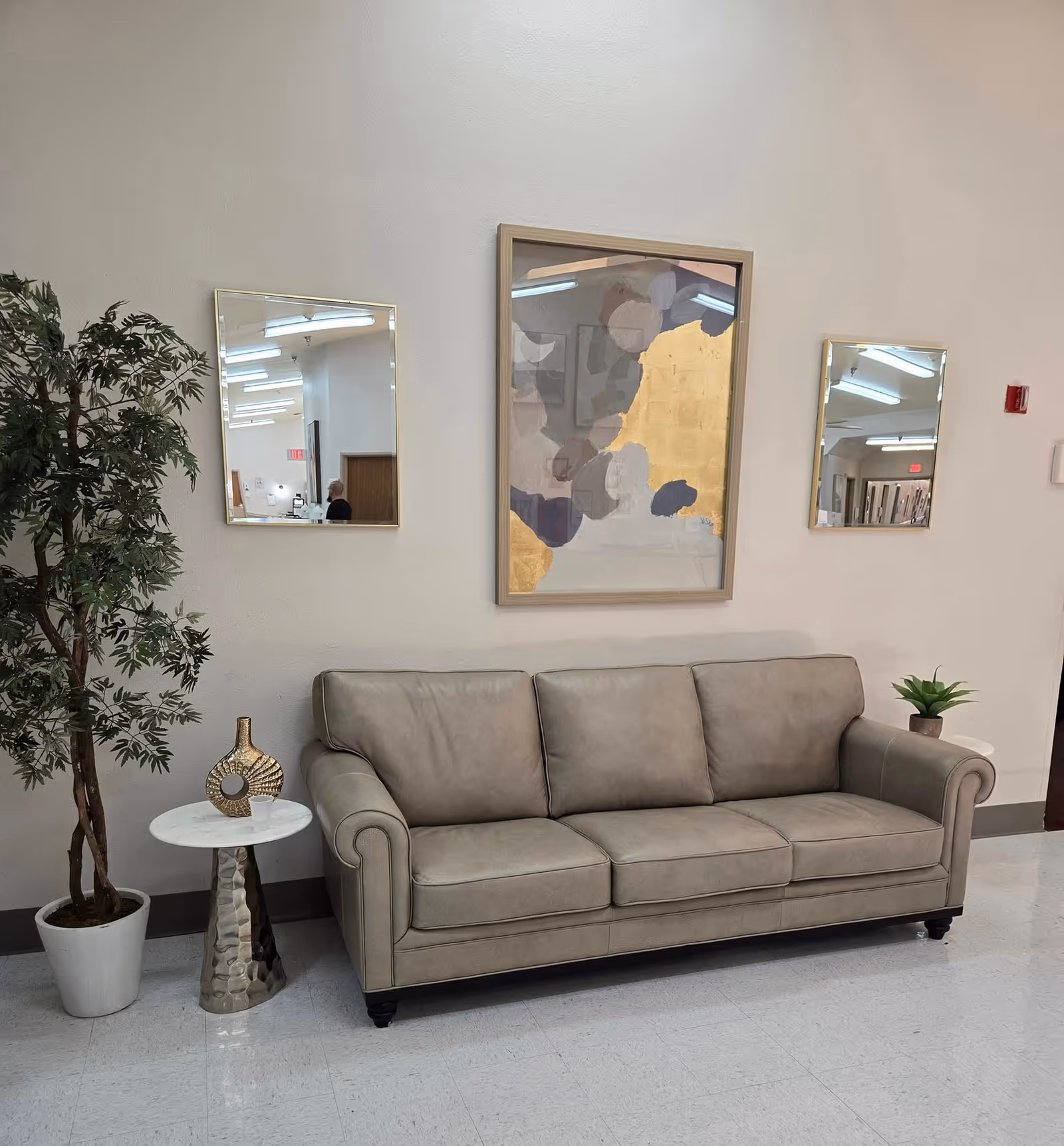 A beige leather sofa placed against a white wall with two square mirrors on either side and an abstract painting with gold and gray tones in the center. To the left of the sofa is a small round side table with a decorative gold vase and a white cup, and a potted artificial tree. To the right of the sofa is a small white side table with a small green plant. The floor is light-colored tile.