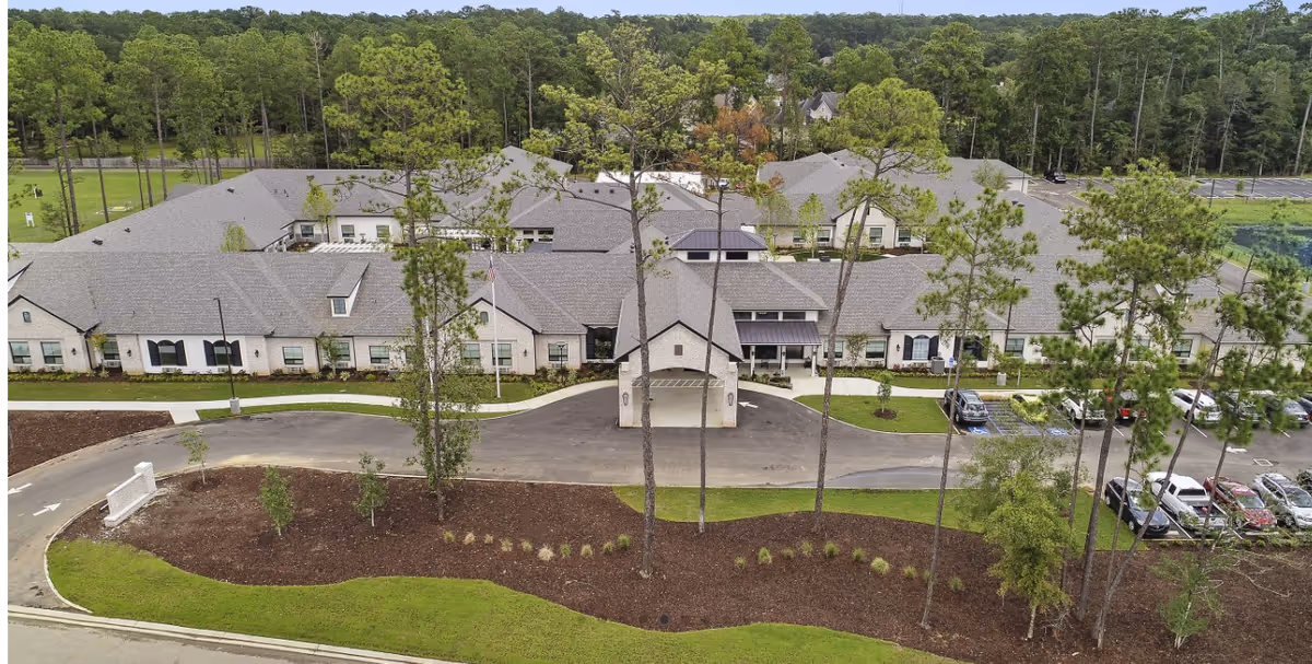 Aerial view of a large single-story senior living facility named Sage Lake, surrounded by trees and greenery. The building has a gray roof and light-colored exterior walls. There is a driveway leading to a covered entrance, with several cars parked in the parking lot to the right. The facility is set in a wooded area with a well-maintained lawn and landscaping in the front.