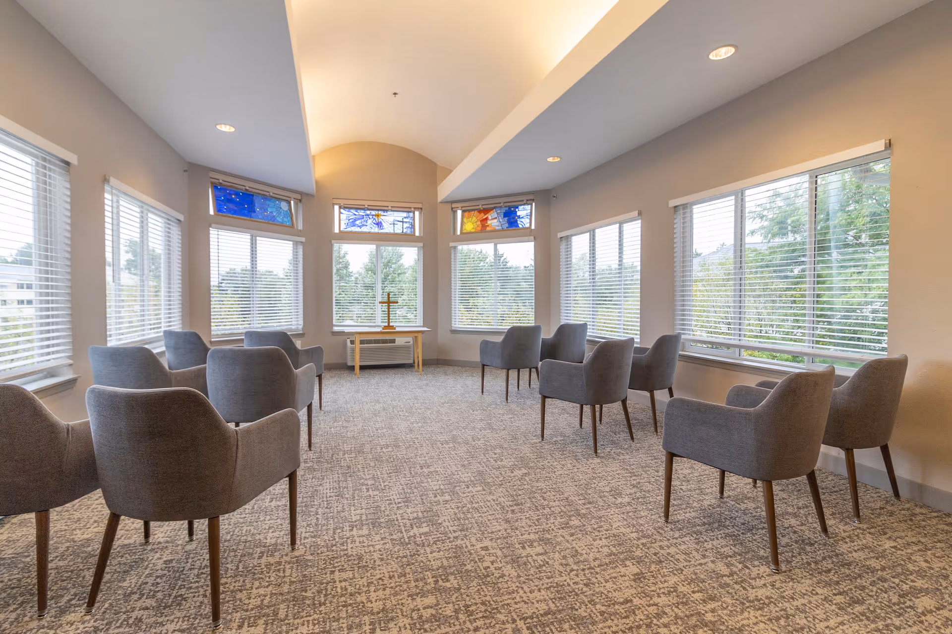 A bright, spacious room with large windows on three sides, featuring gray upholstered chairs arranged in a semi-circle facing a small table with a cross on it. The room has a high, vaulted ceiling with recessed lighting and stained glass panels above the windows.