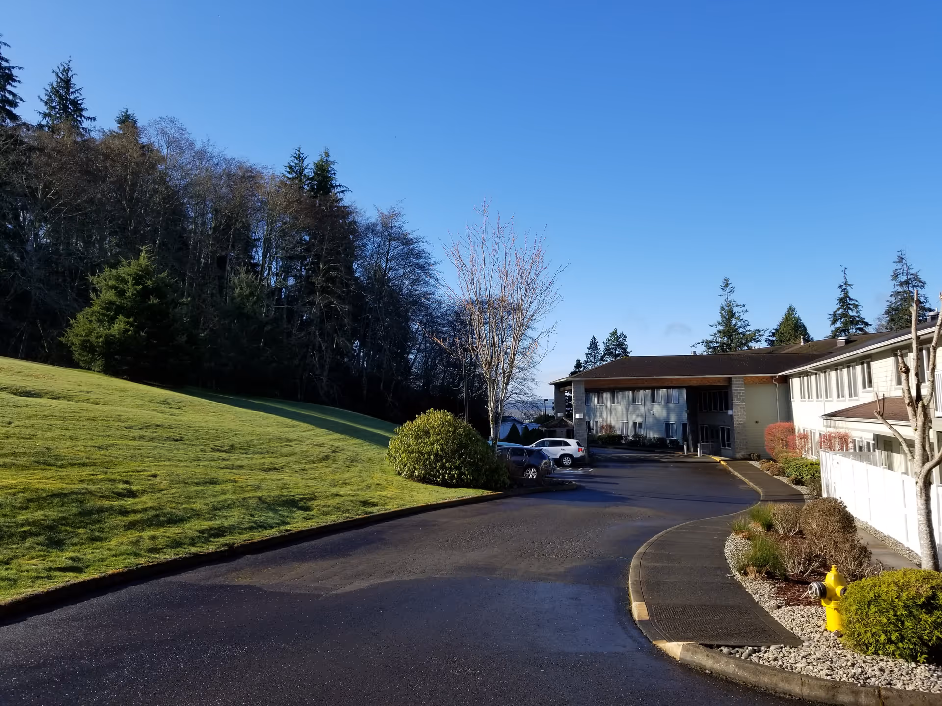 View of the exterior of Westhaven Villa Assisted Living facility on a clear day, showing a paved driveway, parked cars, landscaped bushes, a grassy hill, and trees in the background.