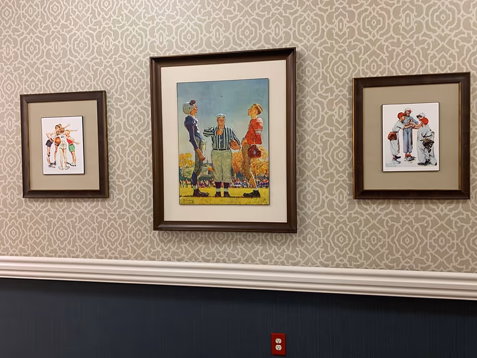 Three framed paintings hanging on a patterned wallpapered wall above a white chair rail. The paintings depict vintage-style illustrations of children and a referee in sports settings. Below the chair rail, the wall is painted dark blue with a red electrical outlet visible.