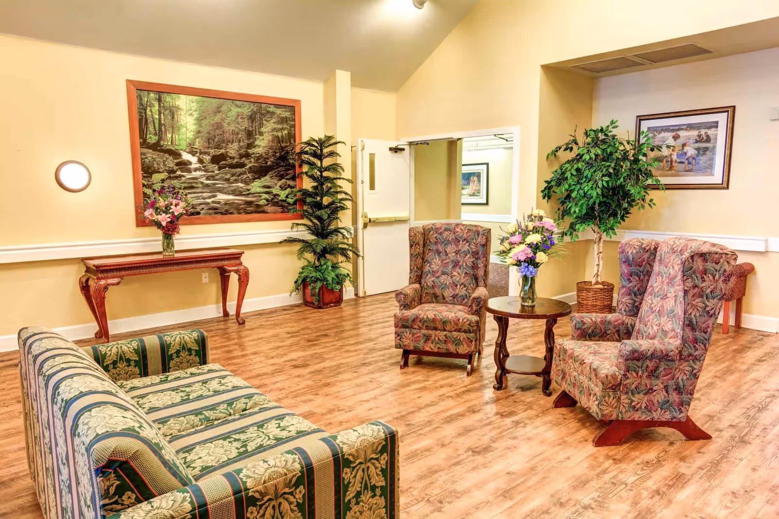 A cozy sitting area in a senior living facility with a patterned green and beige sofa, two floral upholstered armchairs, a round wooden table with a vase of flowers, a wooden console table with a vase of flowers, potted plants, and framed nature-themed artwork on the walls.