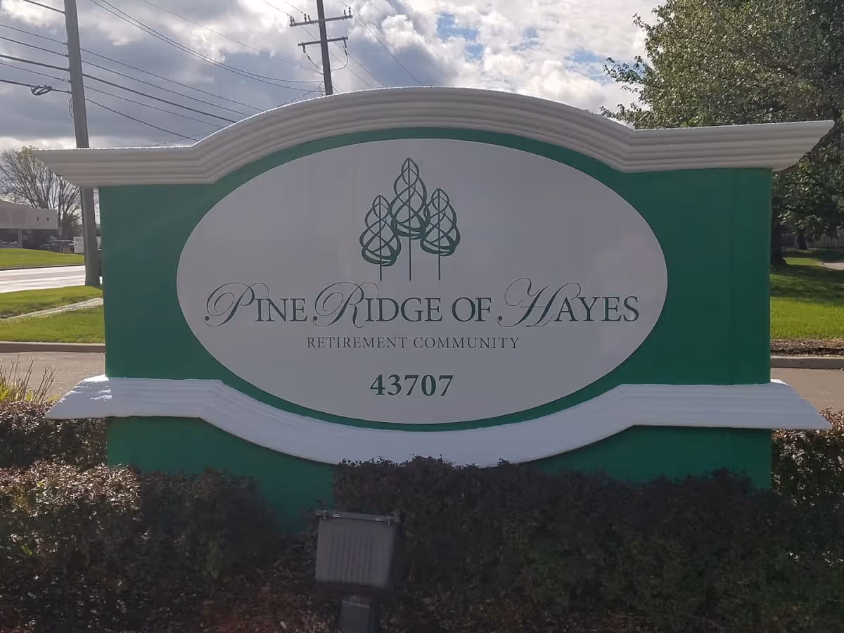 Green and white entrance sign reading 'Pine Ridge of Hayes Retirement Community' with the address 43707 in front of shrubs and a street.