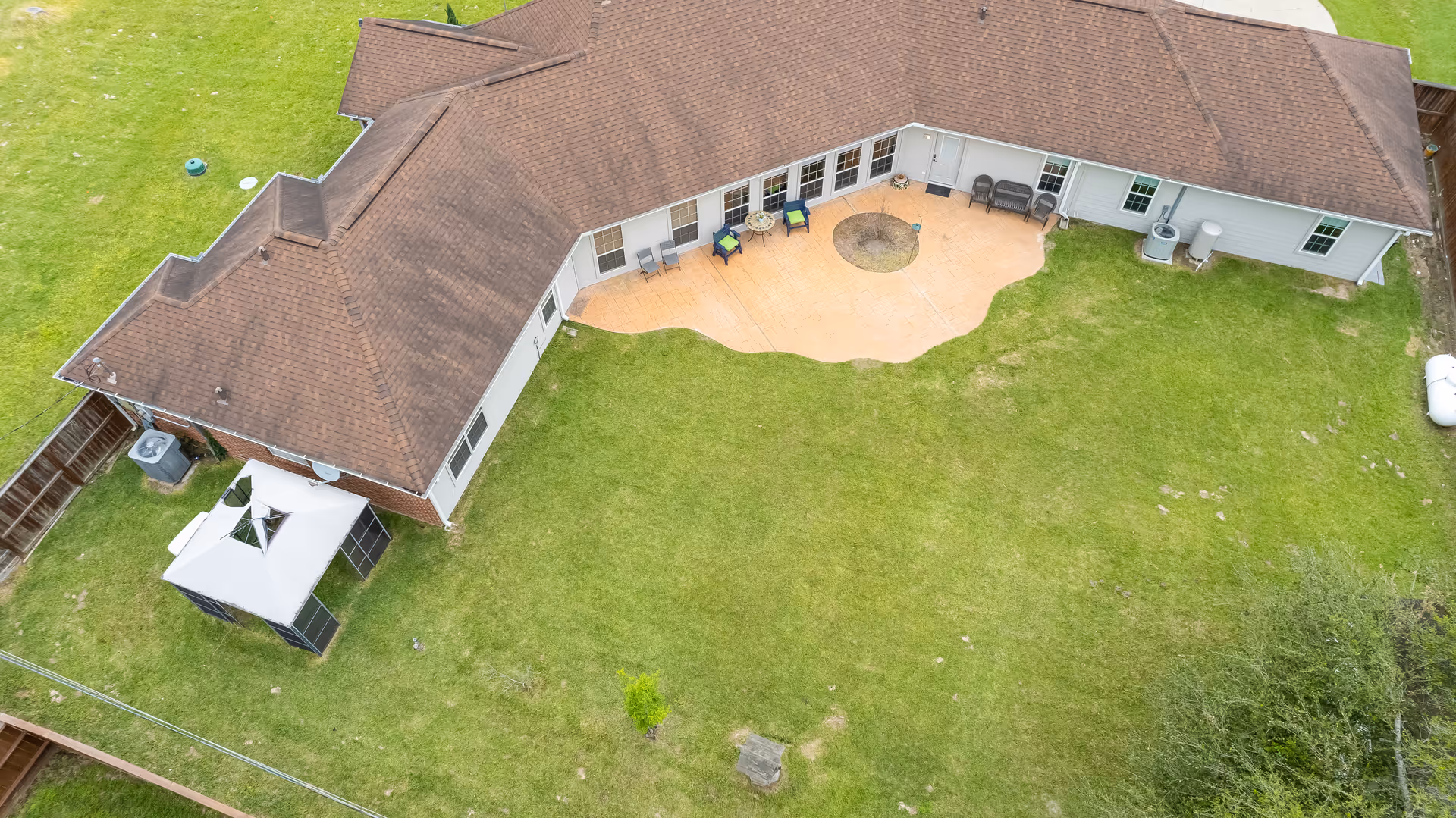 Aerial view of a single-story building with a brown roof and a large green lawn. The building has a curved patio area with outdoor seating and a fire pit in the center. There is a small gazebo structure on the left side of the building and some outdoor equipment near the right side.
