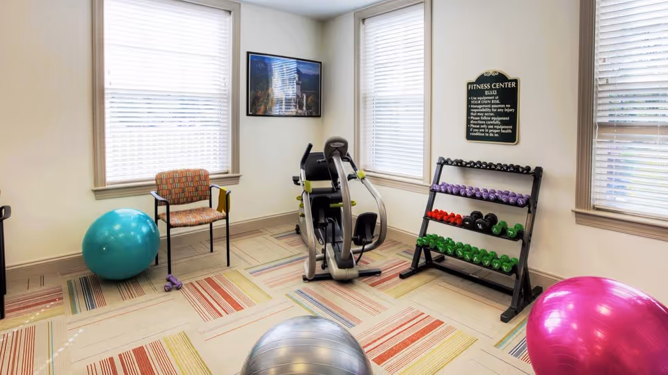 A small fitness center room with exercise equipment including a recumbent bike, a rack of colorful dumbbells, three large exercise balls in teal, silver, and pink, and a chair with patterned upholstery. The room has three windows with blinds and a sign on the wall with fitness center rules.