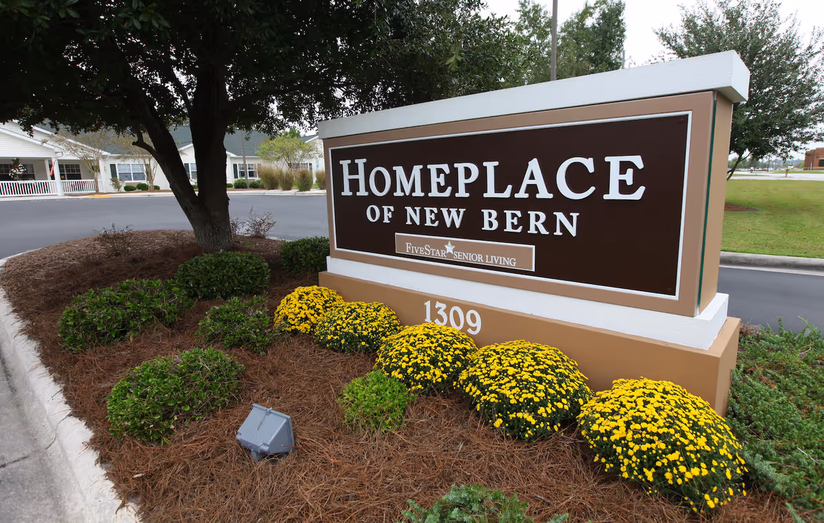 Outdoor view of the entrance sign for Home Place of New Bern, a senior living facility, surrounded by landscaped bushes and yellow flowers with a tree and building in the background.