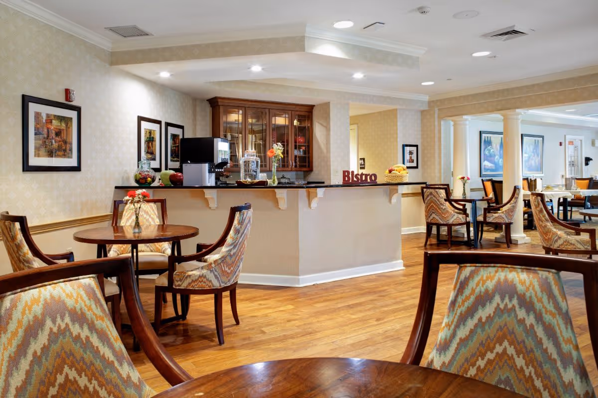 A cozy dining area in a senior living facility featuring round wooden tables with patterned upholstered chairs. In the background, there is a counter labeled 'Bistro' with a coffee machine, glass jars, and a basket of fruit. The room has warm lighting, framed artwork on the walls, and wooden flooring.