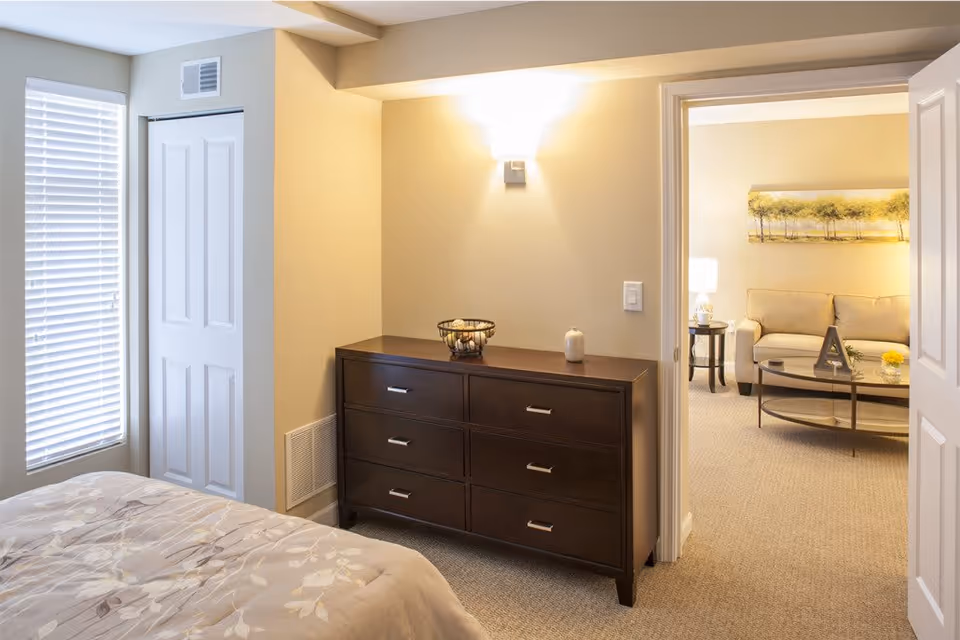 A bedroom with a bed partially visible on the left, a window with blinds, a white closet door, and a dark wooden dresser with six drawers. The dresser has a decorative bowl and a candle on top. Through an open doorway, a living room is visible with a beige sofa, a glass coffee table, a side table with a lamp, and a wall painting of trees.