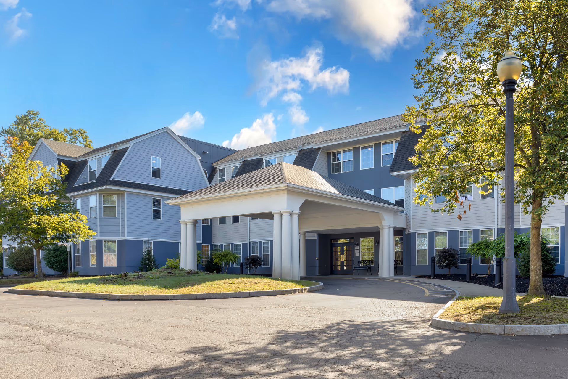 Three-story senior living building front with a covered porte-cochère entrance, surrounding trees, and a blue sky.