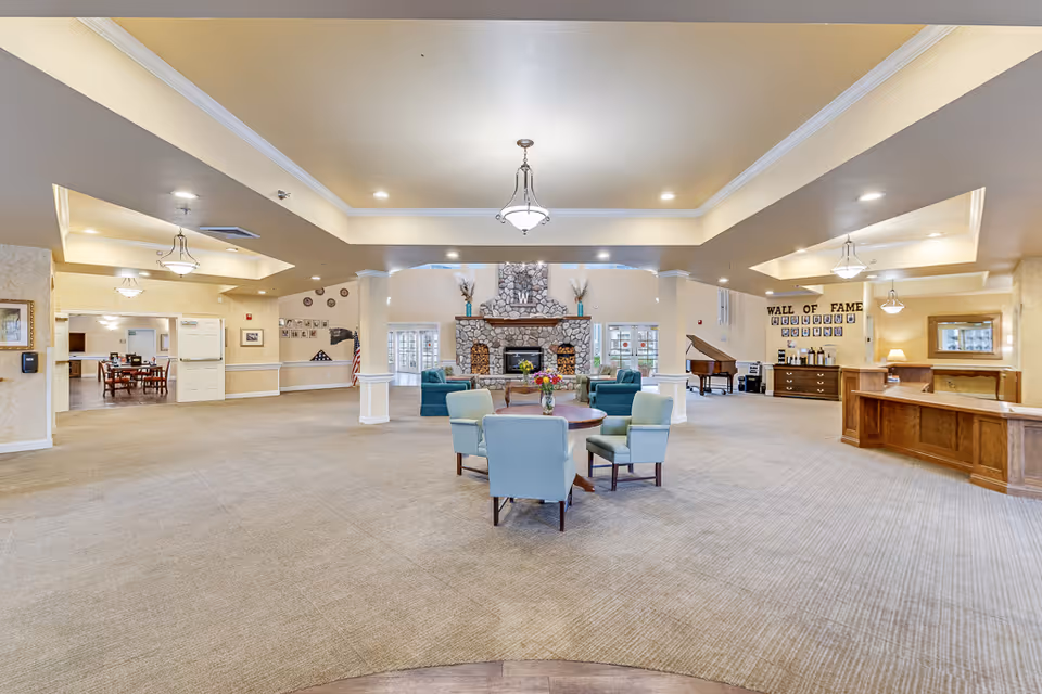 Spacious senior living facility common area with beige carpet and walls, featuring a stone fireplace in the center background flanked by windows. There are several seating arrangements with light blue and green chairs around wooden tables. To the right, there is a wooden reception desk and a wall labeled 'Wall of Fame' with framed photos. A grand piano is also visible near the wall. The ceiling has recessed lighting and hanging light fixtures.