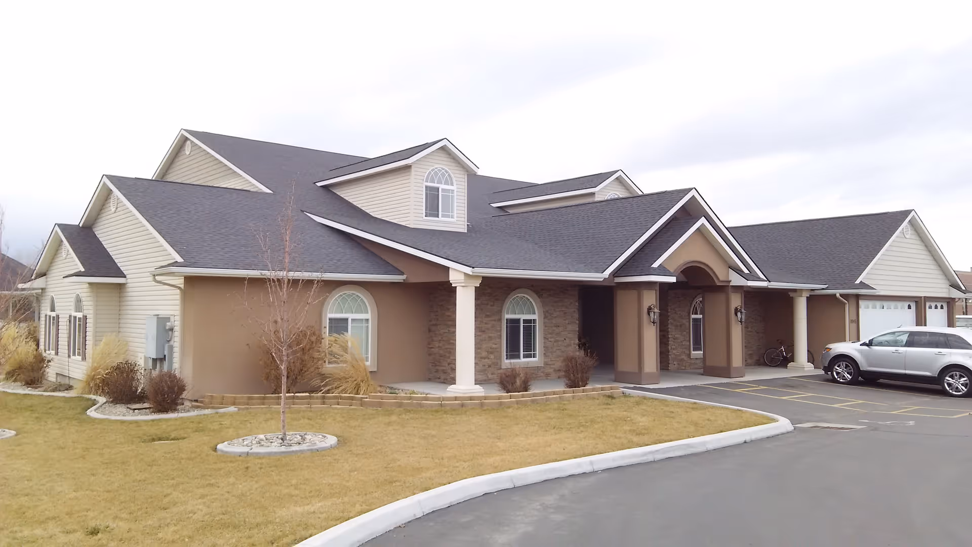 Exterior view of a single-story assisted living facility building with beige siding and stone accents. The building has multiple gabled roofs with dark shingles, arched windows, and a covered entrance supported by white columns. There is a small landscaped area with grass, bushes, and a young tree in front, and a silver SUV parked in the driveway.