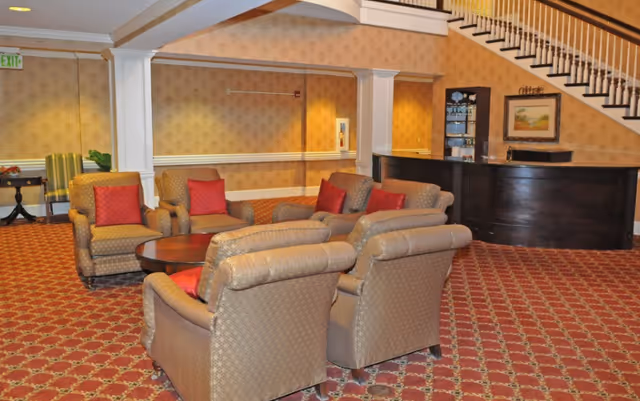 A cozy seating area in a senior living facility with four upholstered armchairs arranged around a round wooden coffee table. The chairs have a patterned fabric and red accent pillows. The room features patterned carpet, wallpaper with a subtle design, white columns, and a dark wooden reception desk under a staircase. A framed painting and a glass display cabinet are visible behind the desk.
