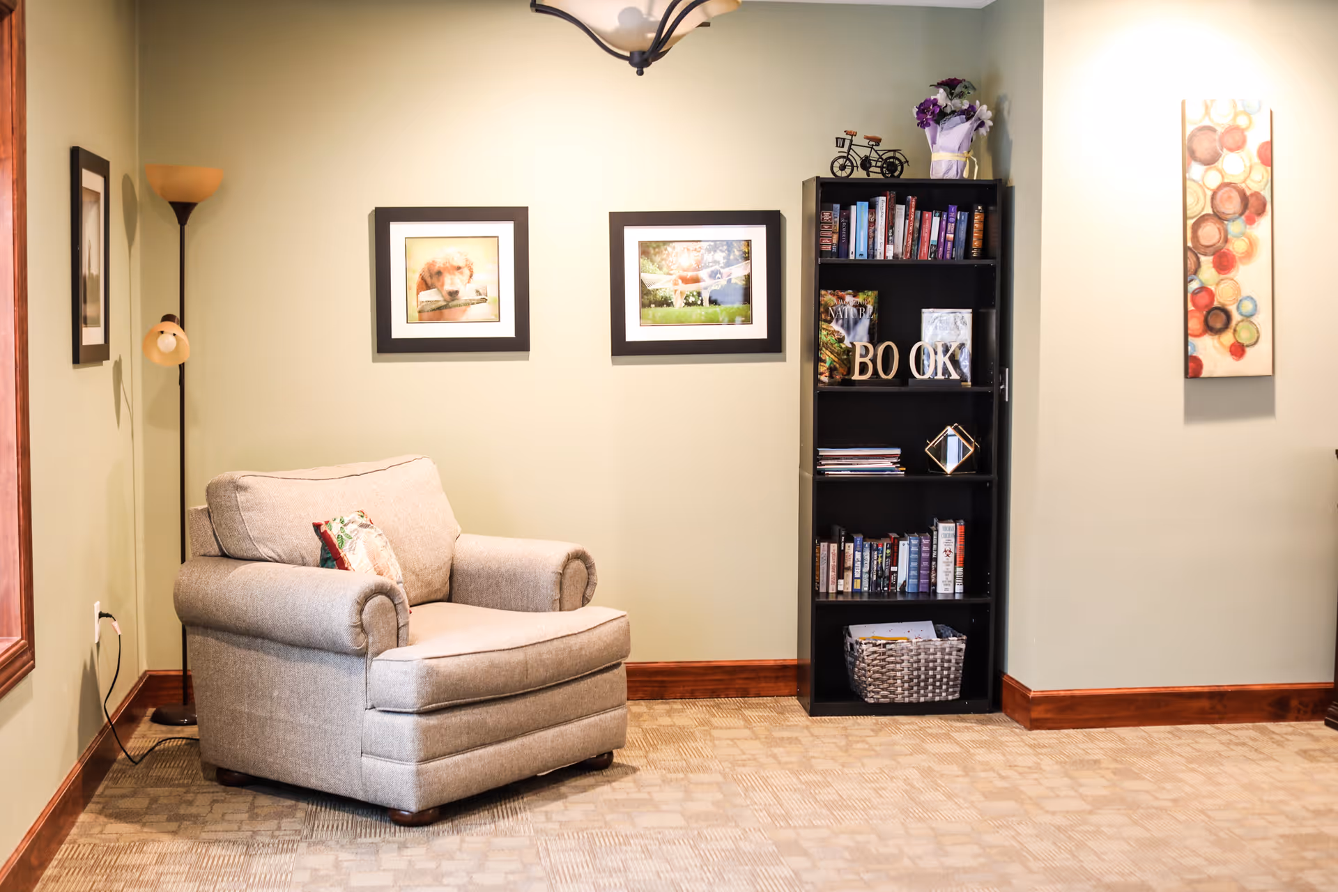 A cozy living room area with a beige armchair, a tall floor lamp, two framed pictures on a light green wall, and a black bookshelf filled with books and decorative items. The room has carpeted flooring and wood trim along the baseboards.