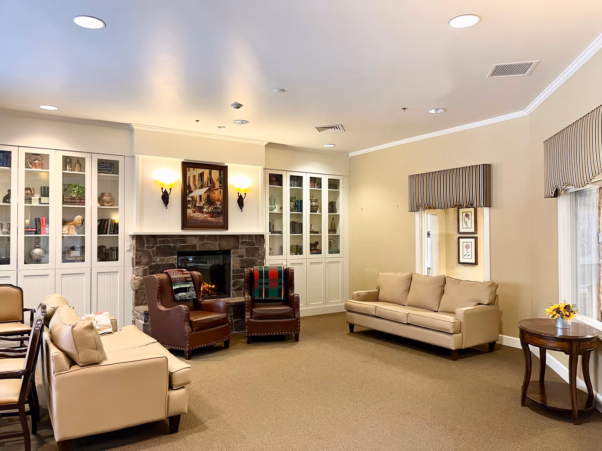 A cozy living room area in an assisted living facility featuring two beige sofas, two brown leather armchairs with patterned blankets, a stone fireplace with a painting above it, built-in white cabinets with glass doors displaying decorative items, and a small round wooden table with a vase of sunflowers near a window with striped valances.