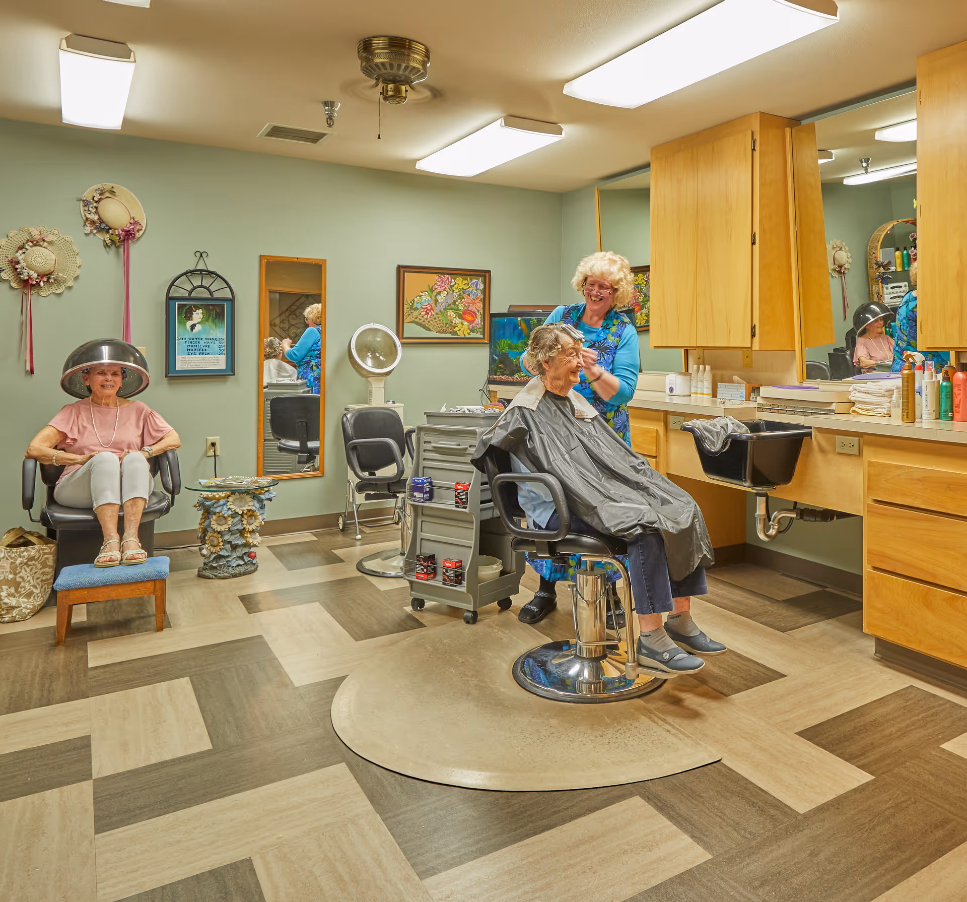 A senior living facility hair salon with two elderly women. One woman is sitting under a hair dryer, smiling, while the other is seated in a salon chair getting her hair styled by a hairdresser. The room has wooden cabinets, a large mirror, and a patterned floor.