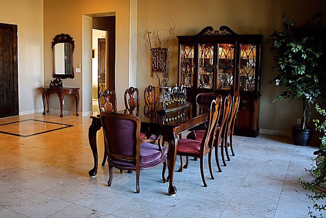 A spacious dining room with a long wooden dining table surrounded by eight chairs with purple upholstery. Behind the table is a large wooden china cabinet with glass doors displaying dishes. To the left, there is a small wooden table with a decorative mirror above it. The floor is tiled with light-colored tiles, and there is a large potted plant on the right side near the wall.