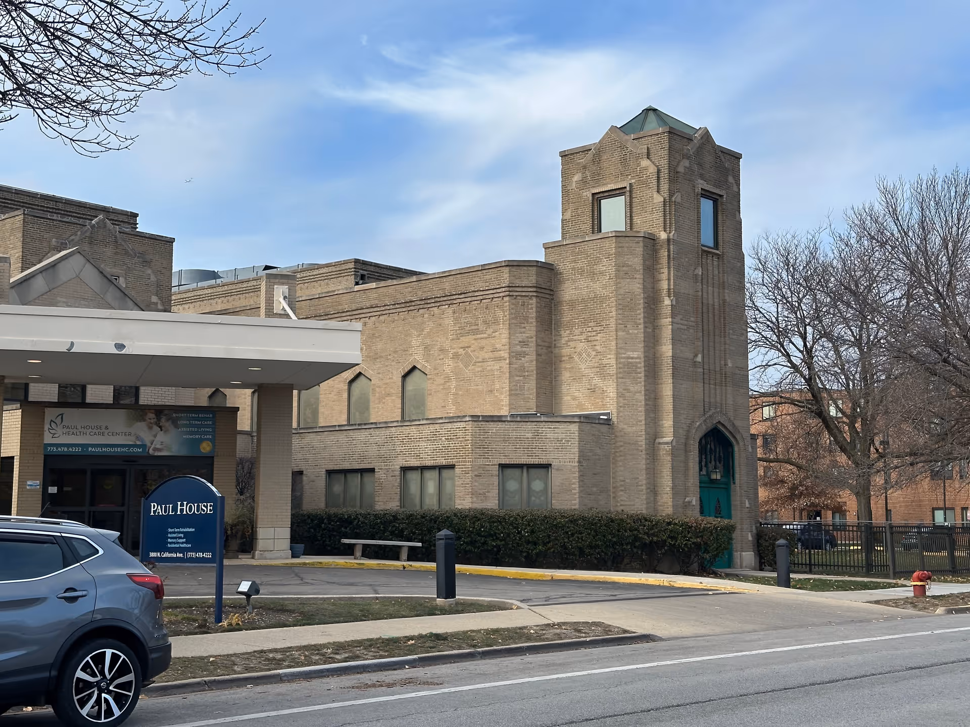 Exterior view of Paul House and Healthcare Center, a beige brick building with a tower-like structure on the right side and a covered entrance on the left. There is a blue sign in front with the facility's name and services listed. A gray car is parked on the street in front of the building, and leafless trees are visible around the property under a partly cloudy sky.