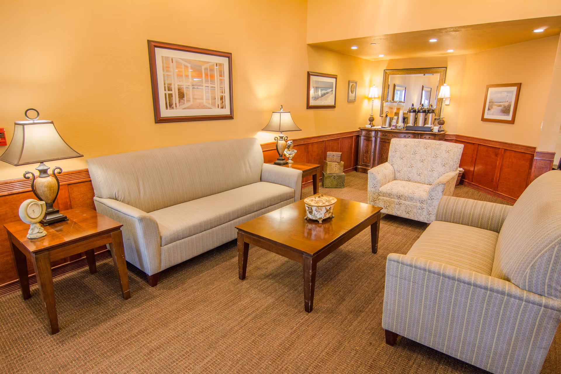 Warmly lit sitting area with two sofas, an armchair, coffee table, side tables and lamps in a communal lounge.