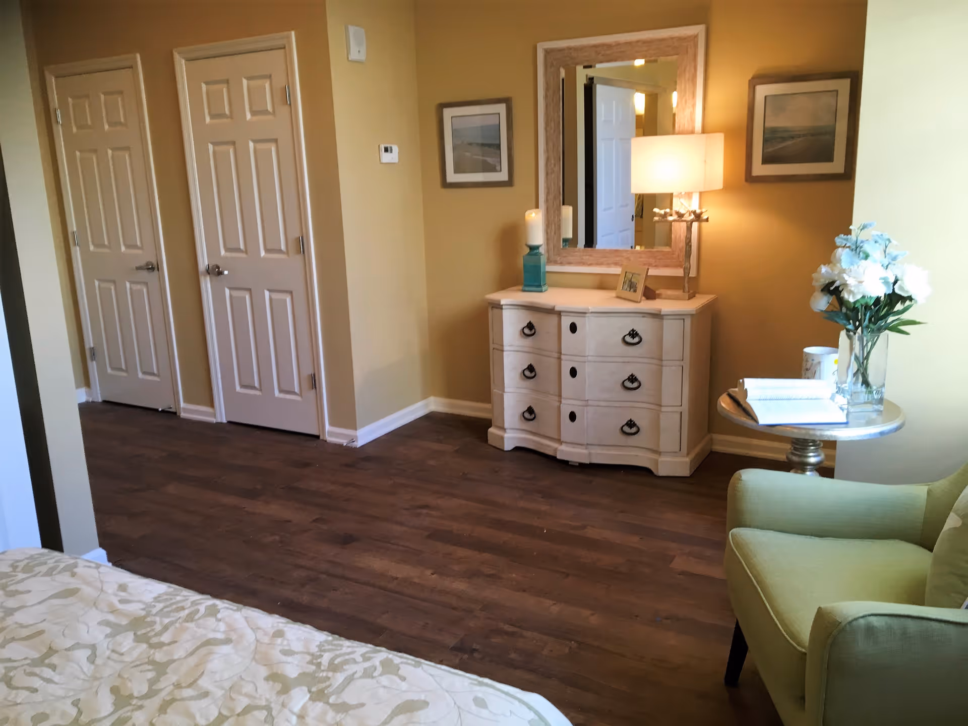 A cozy interior room with wooden flooring, beige walls, and three closed doors. There is a white dresser with a large mirror above it, decorated with a lamp, a candle, and a small framed photo. To the right, there is a green armchair next to a small round table holding a vase of white flowers and an open book. Part of a bed with a patterned bedspread is visible in the foreground.