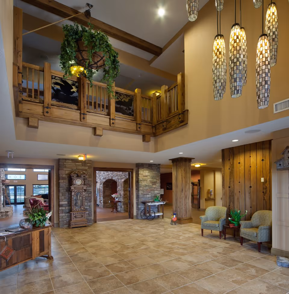 Spacious interior lobby area with tiled floor, wooden beams, and a wooden balcony railing above. The room features hanging mosaic pendant lights, two upholstered armchairs with a small table and plant between them, a wooden grandfather clock, stone archways, and various plants and decorative items.