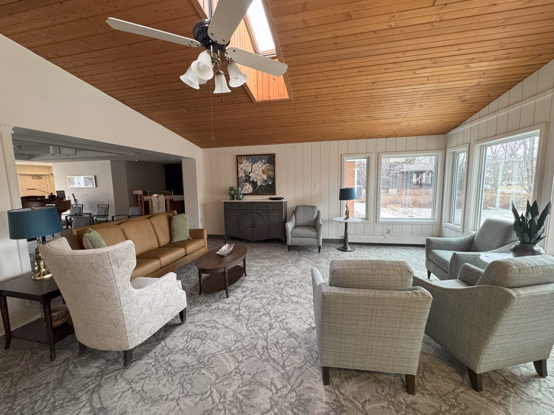 Communal living room with sofas and armchairs arranged around a coffee table, large windows, and a wood-paneled vaulted ceiling with a ceiling fan.