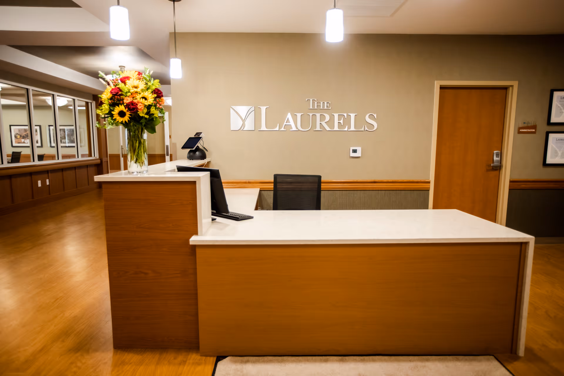 Reception desk area inside The Laurels senior living facility with a large bouquet of flowers on the counter, a computer monitor, and a chair behind the desk. The wall behind the desk displays the sign 'The Laurels'. There is a hallway to the left with framed pictures on the wall and a closed door to the right.
