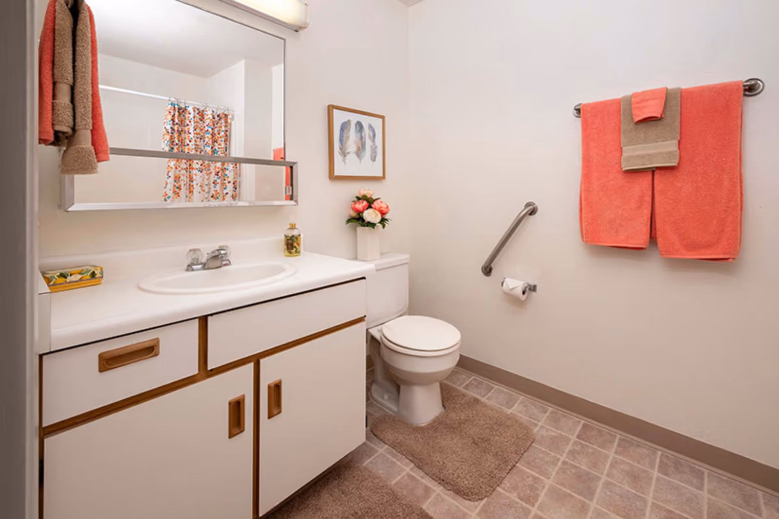 A clean bathroom with a white vanity and sink, a large mirror above the sink, a toilet next to the vanity, and a towel rack on the wall holding coral and beige towels. There is a floral shower curtain visible in the mirror reflection, a small vase with flowers on the toilet tank, and a framed picture of feathers on the wall.