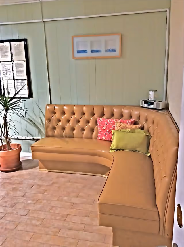 A corner seating area with a tan tufted leather bench adorned with colorful cushions in red, yellow, and green. The walls are painted light green with a framed picture hanging above the bench. A potted plant is placed near a window with black framing on the left side. A small white radio or speaker is positioned on the right corner of the bench.