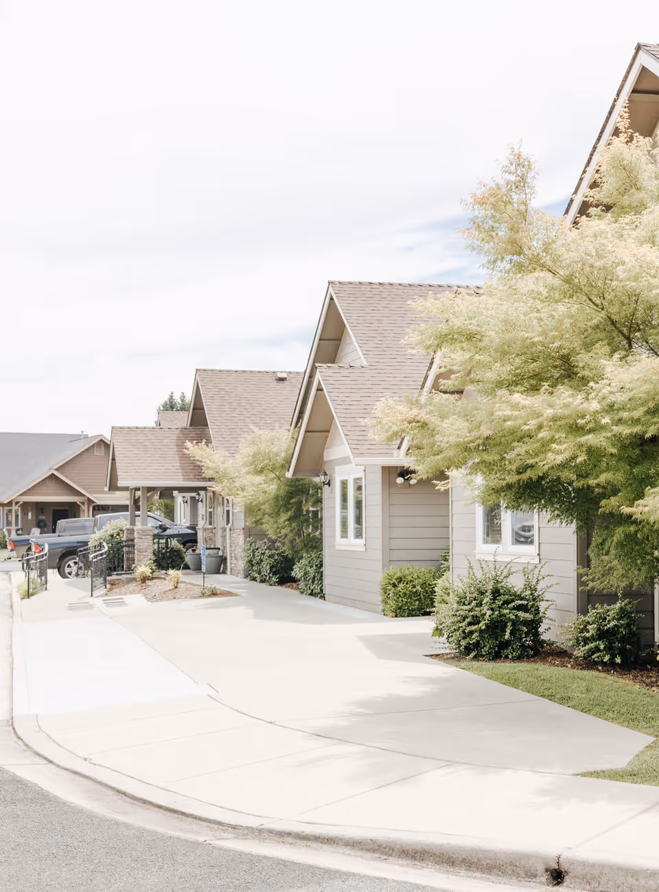 Exterior view of a senior living facility showing a row of small houses with beige siding and brown roofs, surrounded by green bushes and trees, with a concrete sidewalk and driveway in front.
