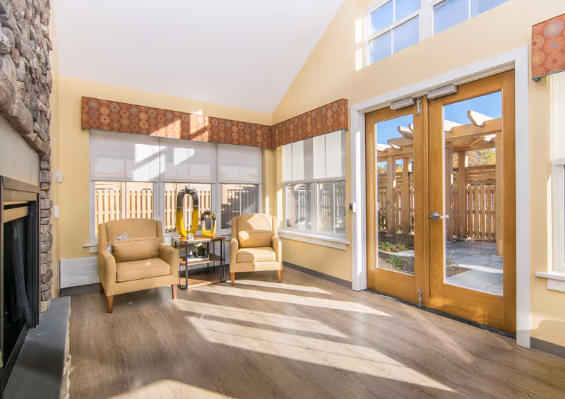 A bright and cozy sitting area with two beige armchairs and a small table between them, featuring decorative glass objects. The room has large windows with patterned valances, a stone fireplace on the left, and wooden double doors leading to an outdoor patio with a wooden pergola.