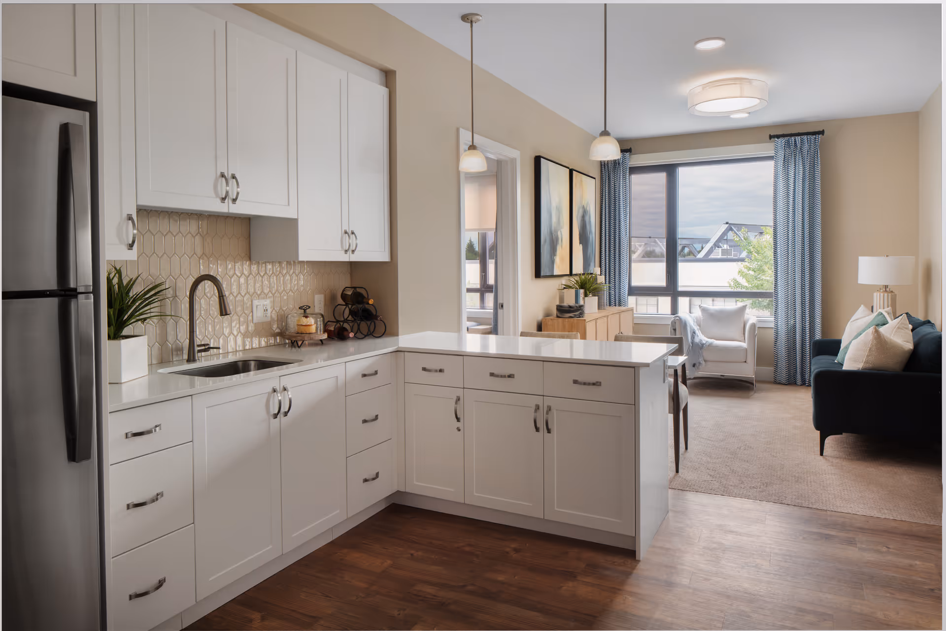 A modern kitchen with white cabinets, a stainless steel refrigerator, and a sink with a faucet. The kitchen opens into a living area with a large window, blue curtains, a white armchair, a dark blue sofa with pillows, a wooden sideboard, and two abstract paintings on the wall.