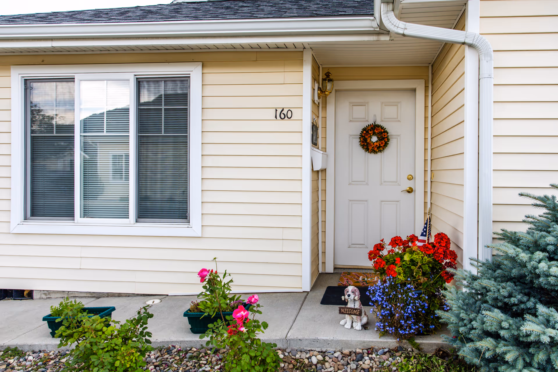 Entrance to a beige siding building with a white door decorated with an autumn wreath. The door is flanked by a small porch area with red and purple flowers and a small dog statue holding a welcome sign. The building number 160 is displayed next to a window with closed blinds.
