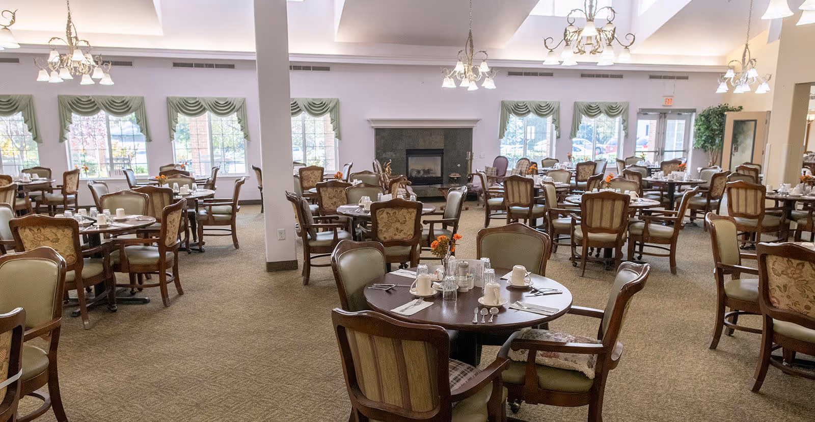 A spacious dining room in a senior living facility with multiple round tables set with cups, glasses, and silverware. The room features upholstered wooden chairs, large windows with green valances, chandeliers hanging from a high ceiling, and a fireplace in the background.
