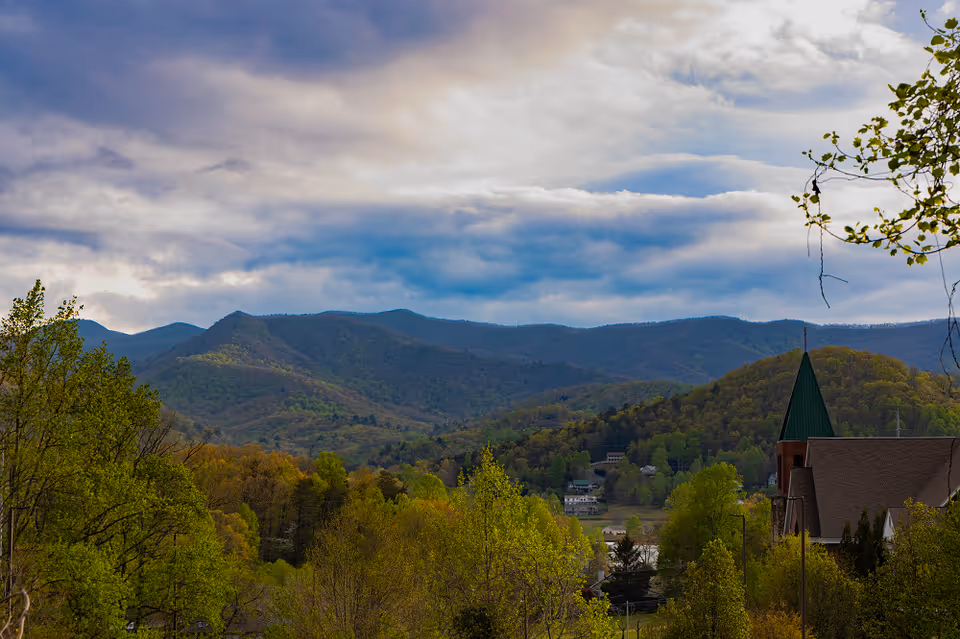 Scenic view of a mountainous landscape with lush green trees in the foreground and a partly cloudy sky. A building with a green pointed roof is visible on the right side.