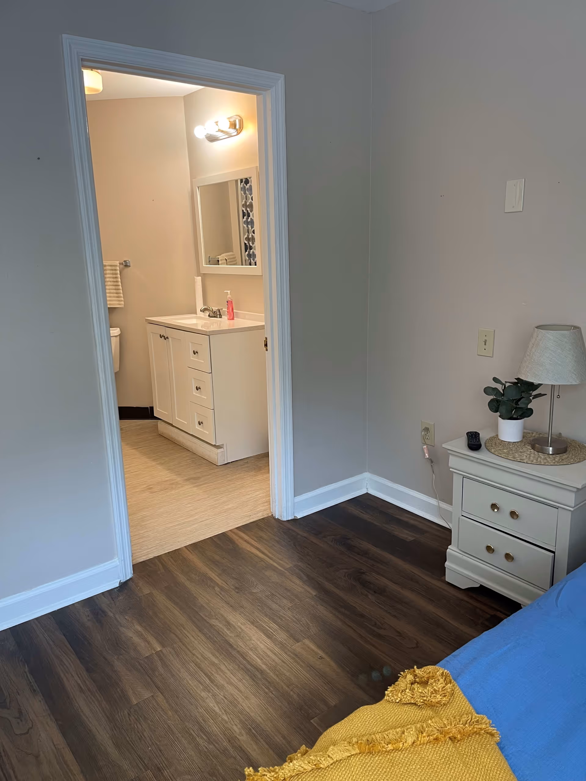 View of a bedroom corner with a nightstand holding a lamp, a small plant, and a remote control. The bed has a blue blanket and a yellow throw. An open door leads to a bathroom with a white vanity, mirror, and light fixture above it.