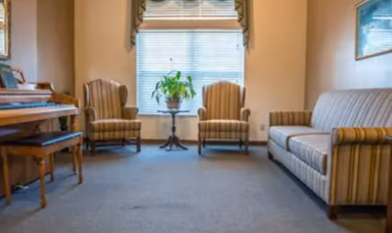 A cozy living room with two striped armchairs and a matching striped sofa arranged around a small round table with a potted plant. A piano with a bench is on the left side near a window with blinds partially open, allowing natural light into the room.