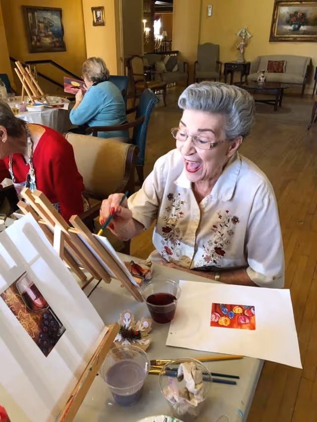 An elderly woman with gray hair and glasses is joyfully painting on a small canvas at a table in a well-lit room. She is wearing a white blouse with floral embroidery. On the table are painting supplies including brushes, cups with water or paint, and a small colorful painting. In the background, other elderly individuals are also engaged in painting activities, seated on chairs in a cozy room with wooden floors and framed artwork on the walls.