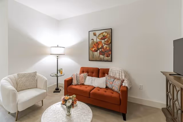 A cozy living room with a burnt orange tufted sofa adorned with multiple decorative pillows and a throw blanket. To the left of the sofa is a white upholstered armchair with a textured pillow. A round white coffee table with a vase of flowers sits in front of the sofa. A floor lamp with a white shade stands behind a small glass side table next to the armchair. On the wall above the sofa is a framed painting of orange flowers. A wooden TV stand with a flat-screen television is visible on the right side of the image.