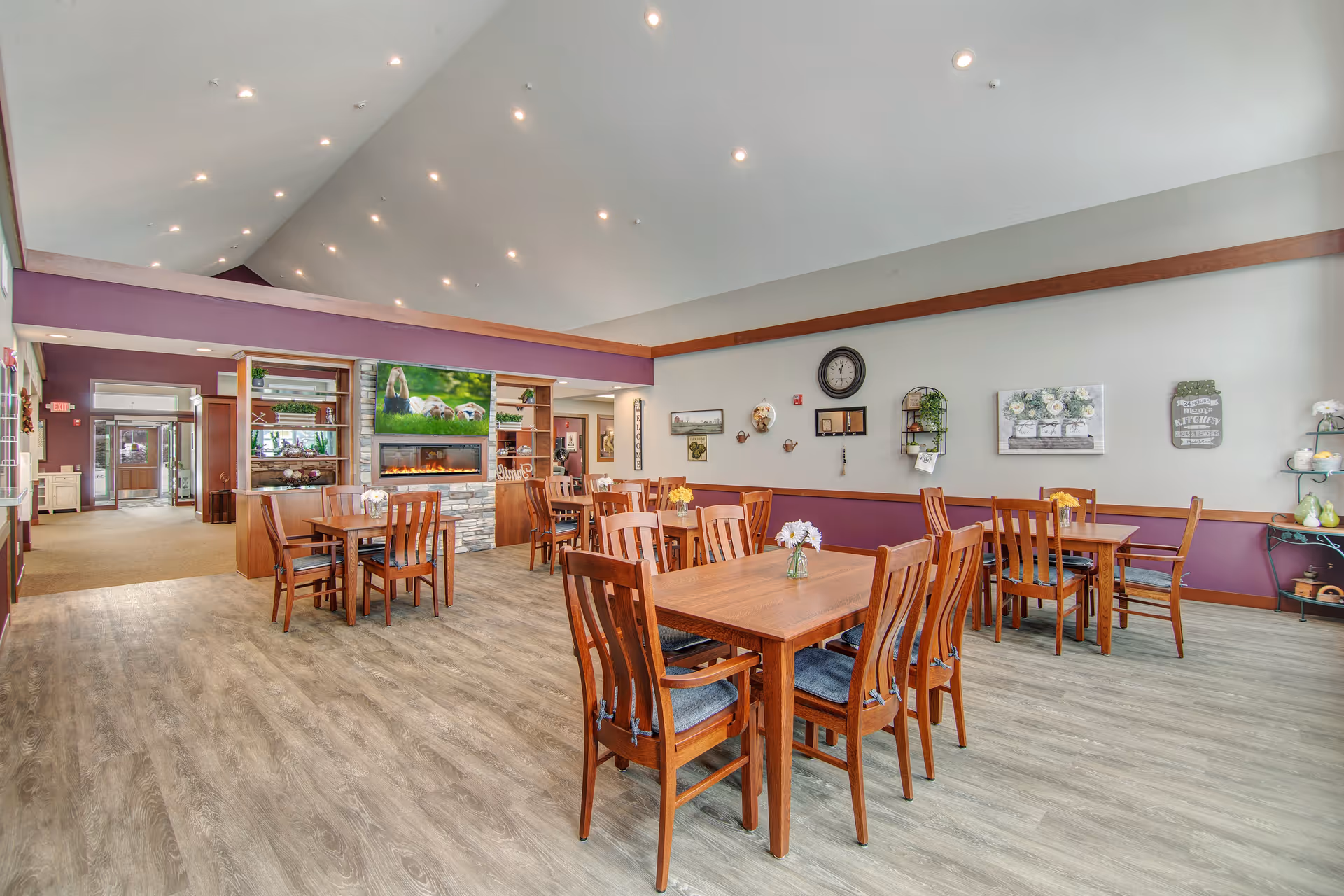 A spacious dining area in a senior living facility with wooden tables and chairs arranged neatly. The room features a high ceiling with recessed lighting, a stone fireplace with a TV above it, and walls decorated with clocks, paintings, and shelves with plants and decorative items. The floor has a wood-like finish, and the color scheme includes purple and neutral tones.