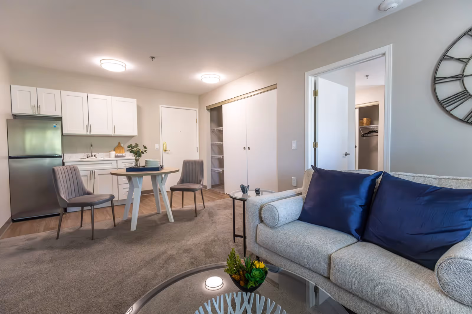 Open-plan living area with a sofa and blue pillows, glass coffee table, round dining table with chairs, and a small kitchenette with stainless steel fridge.