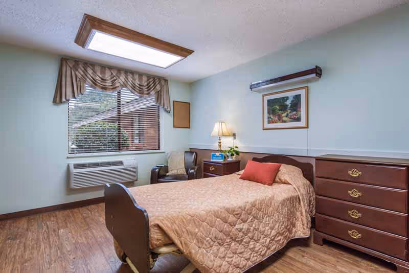 A furnished senior living bedroom with a single bed, bedside table and lamp, dresser, armchair, and a window with blinds.